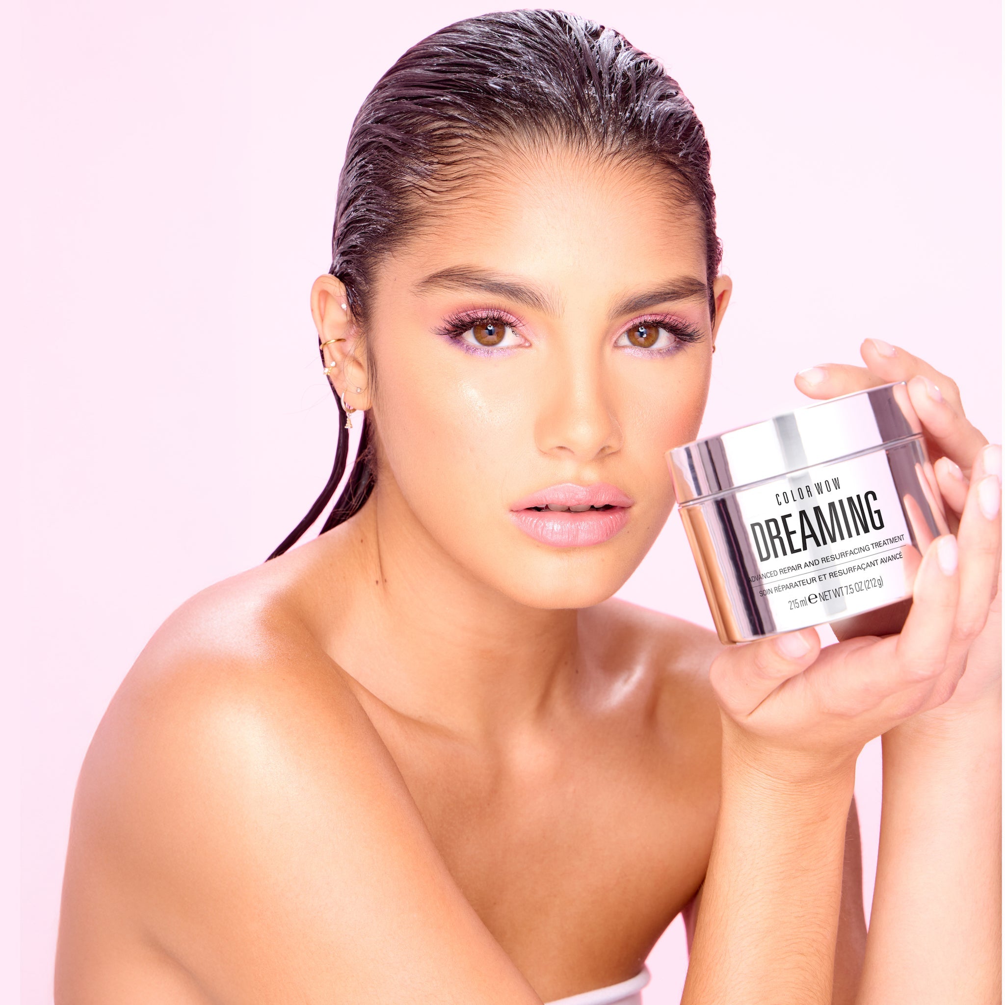A woman with sleek, glowing skin and pink eye makeup holds a jar of Color Wow Dreaming Advanced Repair and Resurfacing Treatment near her face, set against a soft pink background. The silver jar shines, highlighting the product's luxurious and hydrating qualities.